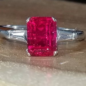 May include: A silver ring with a large, bright red emerald-cut ruby gemstone and two smaller baguette-cut diamonds on either side.