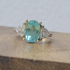 May include: A gold ring featuring a large, oval, light blue gemstone as the centerpiece, flanked by two smaller, triangular, clear gemstones. The ring is set on a wooden surface.