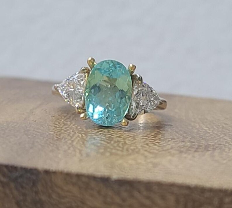 May include: A gold ring featuring a large, oval, light blue gemstone as the centerpiece, flanked by two smaller, triangular, clear gemstones. The ring is set on a wooden surface.
