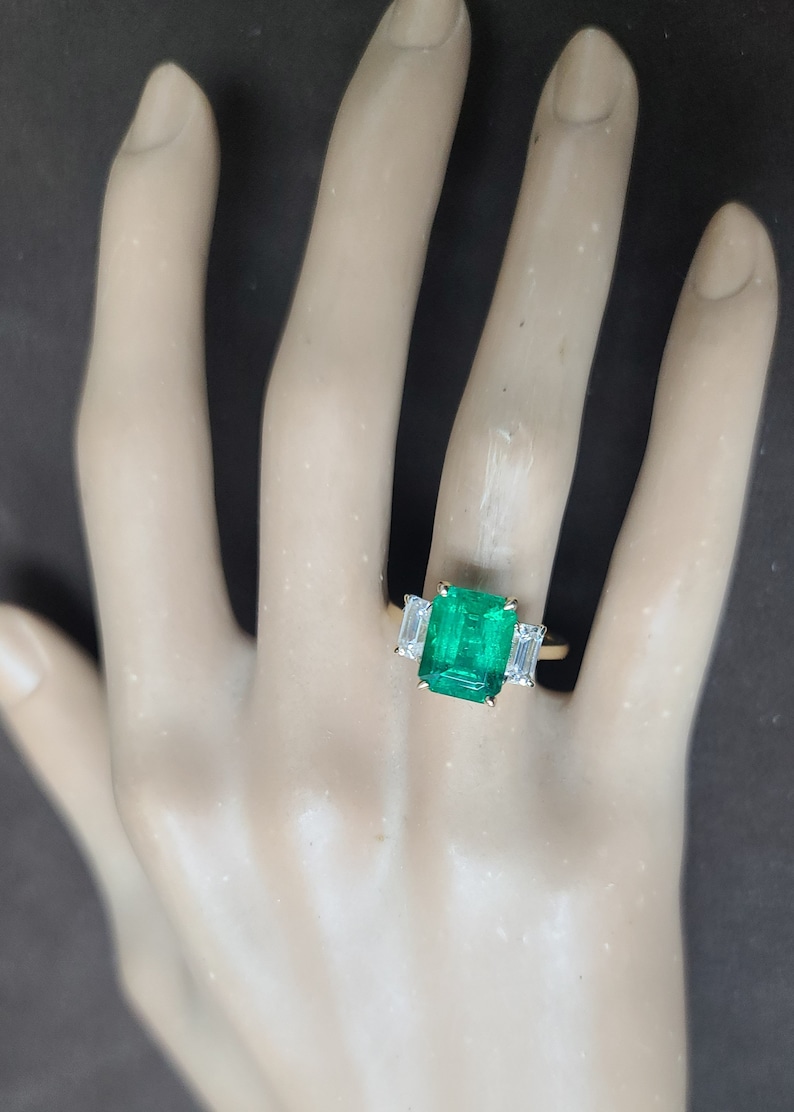 Op de afbeelding: Een gouden ring met een grote smaragdgroene edelsteen en twee kleinere baguette-geslepen diamanten aan weerszijden.