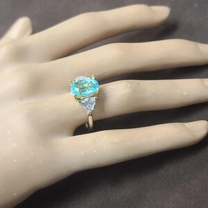 May include: A gold ring featuring a large, oval-cut, light blue gemstone as the centerpiece. Two smaller, triangular, clear gemstones flank the main stone. The ring is set on a neutral-toned background.