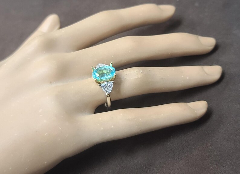 May include: A gold ring featuring a large, oval-cut, light blue gemstone as the centerpiece. Two smaller, triangular, clear gemstones flank the main stone. The ring is set on a neutral-toned background.