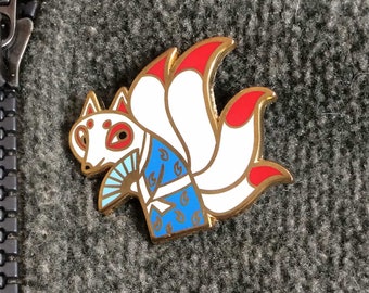 May include: Gold-colored enamel pin featuring a white fox with red accents and a blue and white patterned kimono. The fox has nine tails, a traditional symbol of good luck in Japanese folklore.