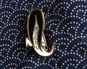 May include: A gold-colored enamel pin featuring a cartoon character with long black hair, a white dress, and a blue eye. The character is in a crouching position with their arms outstretched.