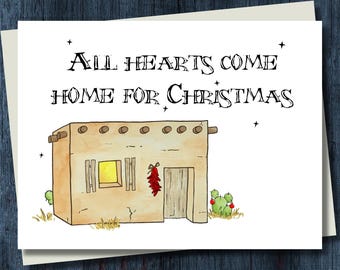 May include: A white greeting card featuring a watercolor illustration of a tan adobe house with a yellow glowing window, a red chili ristra by the door, and green cacti. The text reads: All Hearts Come Home For Christmas with small black stars.