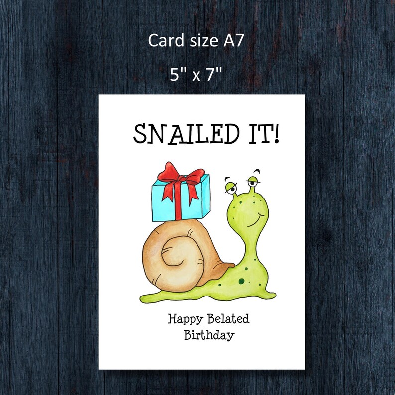 May include: A white greeting card measuring 5 inches by 7 inches featuring a cartoon illustration of a light green snail with a tan shell carrying a light blue gift box with a red ribbon. The text reads SNAILED IT! and Happy Belated Birthday.