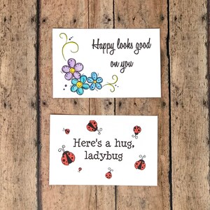 K&ouml;nnte beinhalten: Zwei Gru&szlig;karten mit Blumen- und Marienk&auml;fermotiven. Die obere Karte hat ein Design mit lila und blauen Blumen mit dem Text "Happy looks good on you". Die untere Karte hat ein Design mit einem roten Marienk&auml;fer mit dem Text "Here's a hug, ladybug".