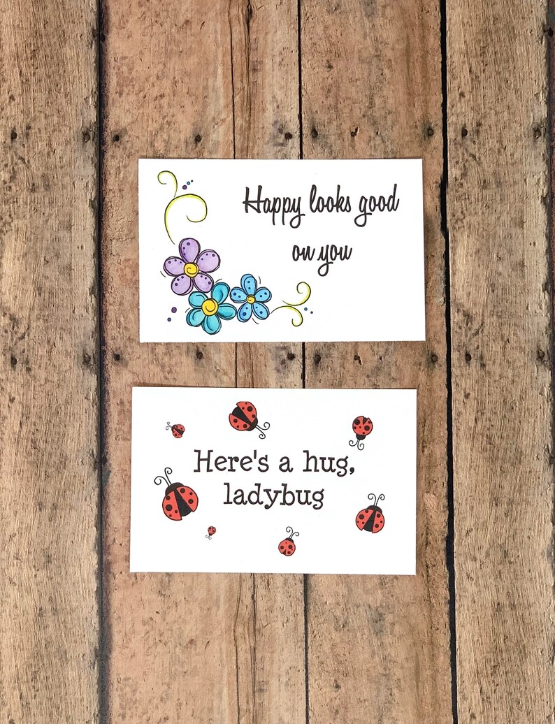 K&ouml;nnte beinhalten: Zwei Gru&szlig;karten mit Blumen- und Marienk&auml;fermotiven. Die obere Karte hat ein Design mit lila und blauen Blumen mit dem Text "Happy looks good on you". Die untere Karte hat ein Design mit einem roten Marienk&auml;fer mit dem Text "Here's a hug, ladybug".