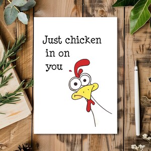 May include: A white greeting card with the text "Just chicken in on you" and a cartoon chicken illustration. The chicken has a red comb and wattle, a yellow beak, and large, expressive eyes. The card is on a wooden surface.