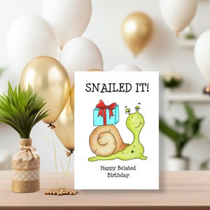 May include: A white rectangular greeting card featuring a cartoon illustration of a light green snail with a brown shell carrying a blue gift box with a red ribbon. The text reads SNAILED IT! and Happy Belated Birthday in black font.