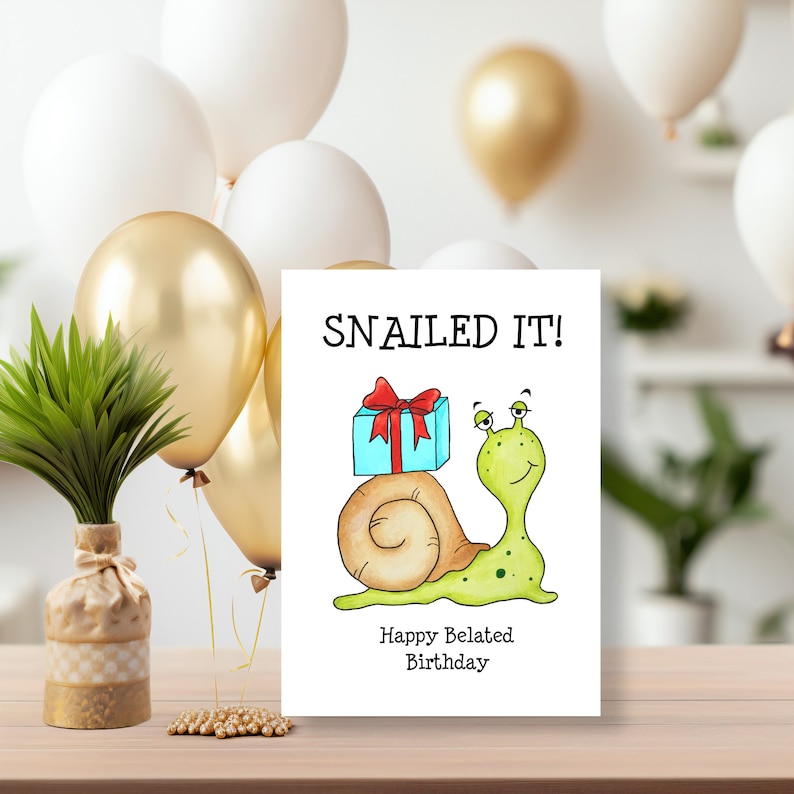 May include: A white rectangular greeting card featuring a cartoon illustration of a light green snail with a brown shell carrying a blue gift box with a red ribbon. The text reads SNAILED IT! and Happy Belated Birthday in black font.