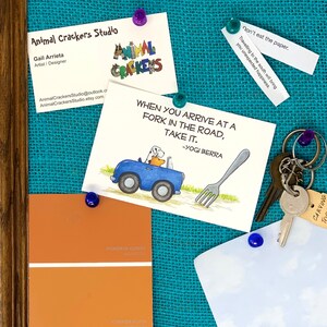 K&ouml;nnte beinhalten: Eine Korktafel mit blauem Hintergrund und einer wei&szlig;en Karte mit einer Cartoon-Illustration eines Hundes, der ein Auto mit einer Gabel auf der Stra&szlig;e f&auml;hrt. Die Karte tr&auml;gt den Text "Wenn du an einer Gabelung ankommst, nimm sie. -Yogi Berra". Es gibt auch zwei Visitenkarten, eine Farbkarte und einen Schl&uuml;ssel mit einem Anh&auml;nger, auf dem "Carving Tan" steht.