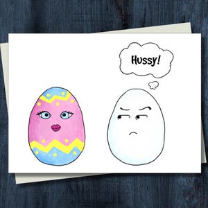 May include: A greeting card featuring a cartoon illustration of two eggs. One egg is decorated with pink, blue, and yellow patterns, eyelashes, and red lips. The other plain white egg has a judgmental expression and a thought bubble saying Hussy!