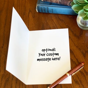 May include: A white greeting card standing open on a wooden surface, featuring the black printed text 'optional: Your custom message here!' in the center. A brown and gold ballpoint pen rests on the surface next to the card.