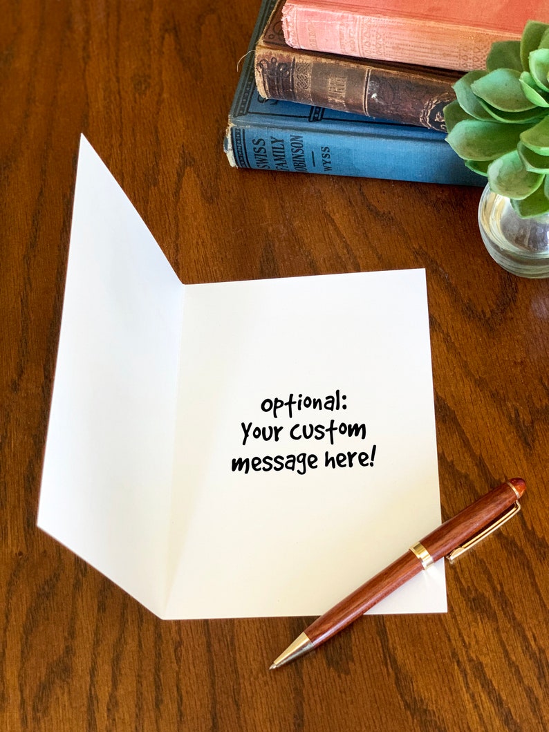 May include: A white greeting card standing open on a wooden surface, featuring the black printed text 'optional: Your custom message here!' in the center. A brown and gold ballpoint pen rests on the surface next to the card.