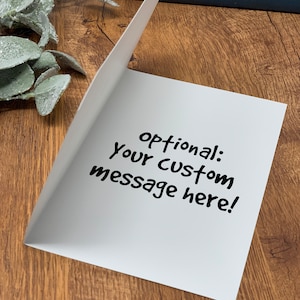 May include: A white greeting card with the text "optional: Your custom message here!" in black. The card is slightly open, revealing the inside. The background is a wooden surface with decorative elements.