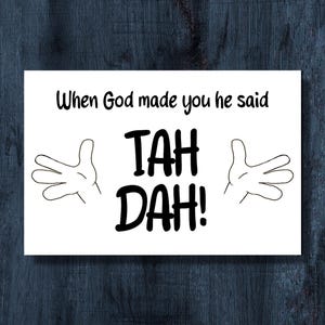 May include: A white rectangular card with the text "When God made you he said TAH DAH!" in black. The card features two cartoon hands with open palms, one on each side of the text. The background is a dark wood grain.