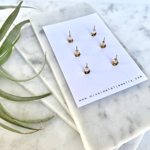 May include: A close-up of the back of a card with four gold earring backs. The card is white with the text "www.minormetaljewelry.com" printed on it.