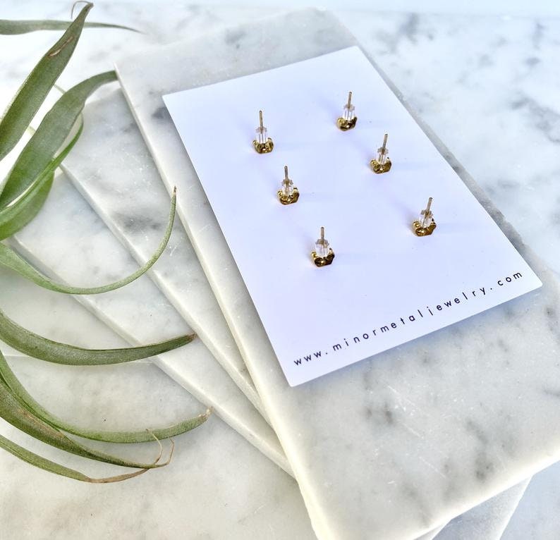 May include: A close-up of the back of a card with four gold earring backs. The card is white with the text "www.minormetaljewelry.com" printed on it.
