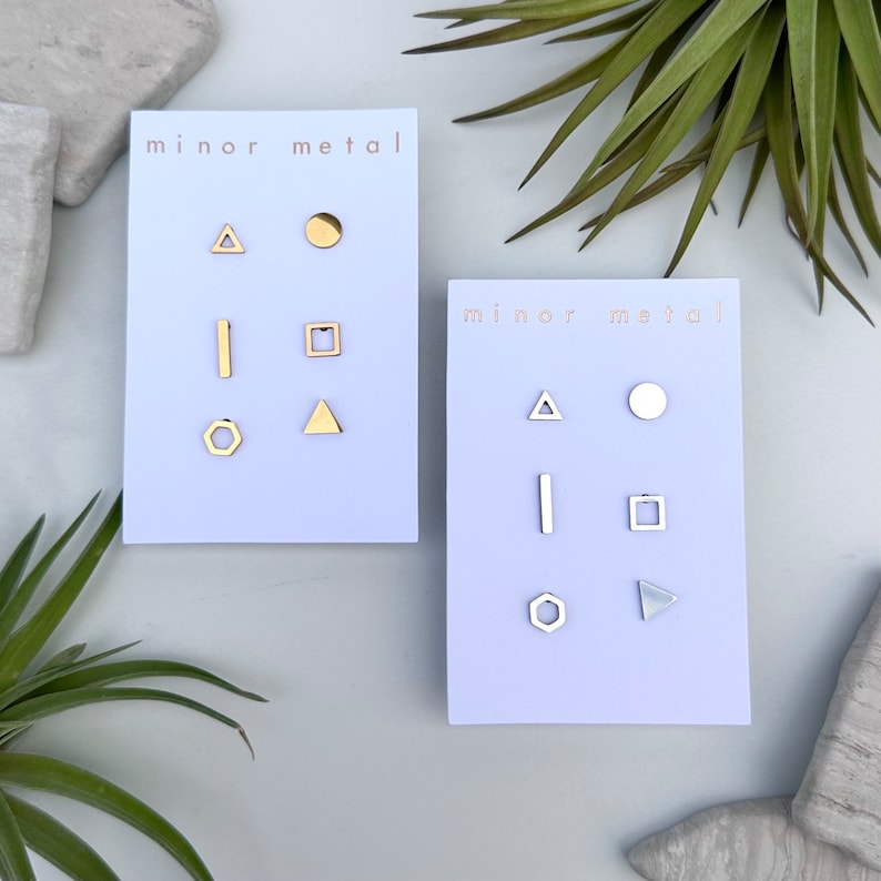 May include: Two sets of geometric stud earrings on white cards. The first set is gold and includes a circle, triangle, square, line, and hexagon. The second set is silver and includes a circle, triangle, square, line, and hexagon. Both cards have the text "minor metal" printed on them.