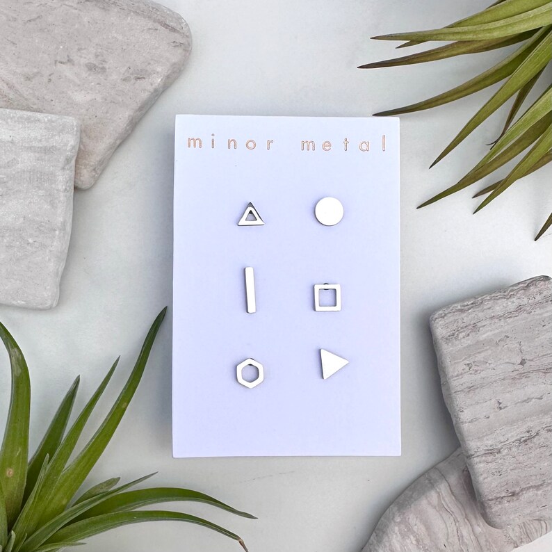 May include: A set of six silver geometric stud earrings on a white card with the text "minor metal". The earrings are a triangle, circle, line, square, hexagon, and a triangle.