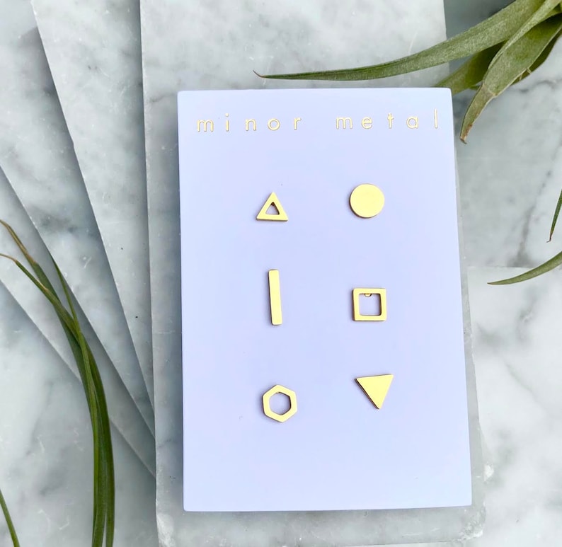 May include: A set of six gold geometric stud earrings on a purple card with the text "minor metal". The earrings are a triangle, circle, line, square, hexagon, and triangle.