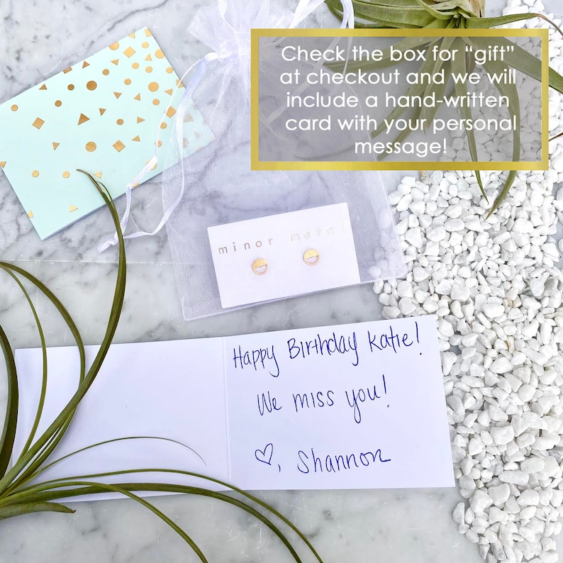 May include: A white card with a handwritten message that says "Happy Birthday Katie! We miss you! Love, Shannon". The card is next to a small white bag with gold earrings inside. The bag has the words "minor muse" printed on it.