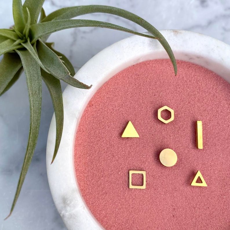 May include: A set of six gold geometric earrings in a white bowl with a pink interior. The earrings are shaped like a circle, square, triangle, hexagon, and two different sized triangles.