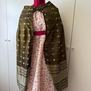 May include: An olive green cape with gold accents and a floral patterned lining. The cape is draped over a dress form, showcasing its design and texture. The lining features a delicate floral print in shades of pink and purple.