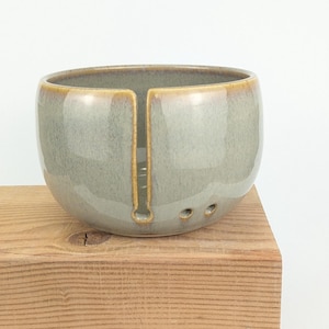 May include: A ceramic yarn bowl in a soft green hue with a golden rim and accents. The bowl has a slot for the yarn and three holes. It is sitting on a wooden block.