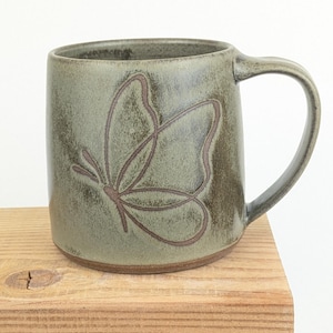 May include: A handmade ceramic mug in a muted green colour, featuring a brown butterfly design. The mug has a rounded shape with a sturdy handle. The butterfly is outlined with brown lines. The mug is sitting on a wooden surface.