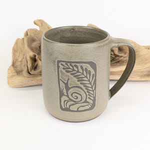 May include: A ceramic mug with a light green glaze and a dark grey handle. The mug features a rectangular design with a snail and leaf illustration. The mug is approximately 10 cm tall and 8 cm in diameter.