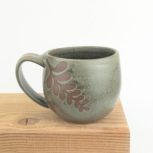 Peut inclure: Mug en céramique fait à la main, avec un glaçage vert-gris et un motif de feuille marron. La tasse a une forme arrondie et une grande anse. Posée sur une surface en bois.