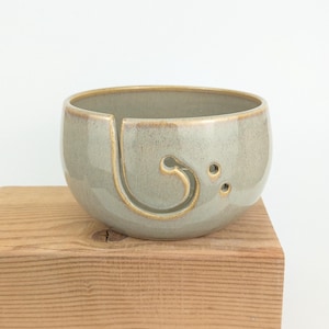 May include: A ceramic yarn bowl in a soft green-grey glaze, featuring a spiral yarn guide and three small holes. The bowl has a rounded shape with a slightly flared rim and sits on a wooden block.