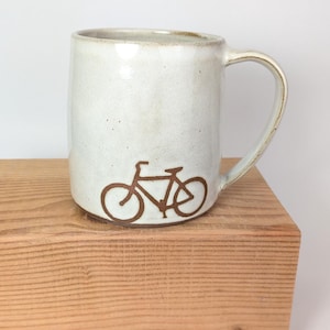 May include: Cream ceramic mug featuring a brown bicycle design. The mug has a cylindrical form with a curved handle. The bicycle graphic is near the base. The mug is placed on a wooden surface. The mug is approximately 10 cm tall.