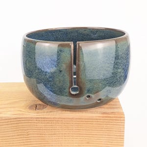 May include: A handcrafted ceramic yarn bowl in shades of blue and green. The bowl has a glossy finish and a slot for yarn to feed through, with three small holes on the side. It sits on a wooden surface.