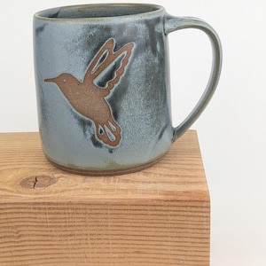 May include: A ceramic mug with a blue-grey glaze and a brown hummingbird design. The mug has a curved handle and sits on a wooden block. The hummingbird is depicted in flight, with its wings spread.