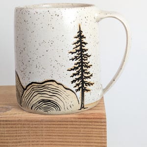 May include: A ceramic mug with a speckled off-white glaze featuring a black line drawing of a tall pine tree and a series of concentric circular lines representing a cross-section of a tree trunk near the base of the vessel.