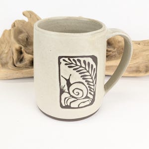 May include: A beige ceramic mug featuring a dark brown illustration of a snail and fern. The mug has a rounded body, a handle, and a dark brown base. The design is within a rectangular frame.