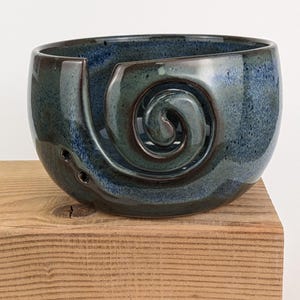 May include: A ceramic yarn bowl in shades of blue and green, with a spiral design. The bowl has a glossy finish and sits on a wooden surface. It has three small holes near the base.