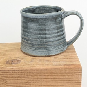 May include: A handmade ceramic mug with a blue-grey glaze. The mug has a cylindrical shape with a rounded handle and horizontal textured lines. It sits on a wooden surface against a white background. The mug is approximately 10 cm tall.
