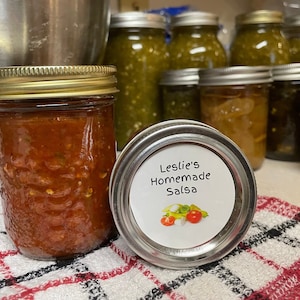 May include: A jar of Leslie's Homemade Salsa with a red and chunky texture, next to a jar lid with a label that reads "Leslie's Homemade Salsa" and a graphic of tomatoes, onions, and peppers. Other jars of salsa are in the background.