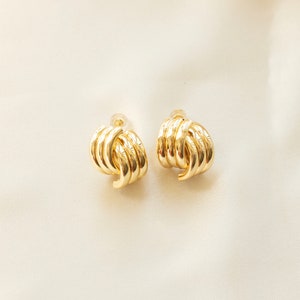 May include: A pair of gold-colored earrings. Each earring features a looped design with multiple curved, parallel bars. The earrings are set against a soft, off-white background, highlighting their metallic sheen and intricate form. The earrings are a classic jewelry item.