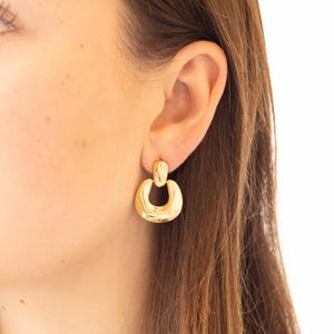 May include: Gold-colored, chunky hoop earrings. The earrings have a rounded, U-shaped design with a small, rectangular post at the top. The earrings are shiny and reflect light.