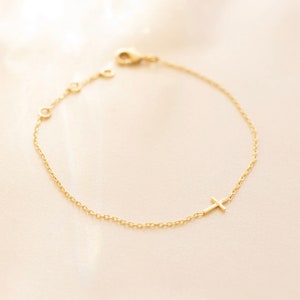 May include: A delicate gold chain bracelet with a small cross charm. The bracelet is made of a fine chain and has a simple, elegant design.