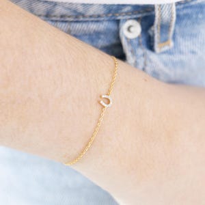 May include: A delicate gold chain bracelet featuring a small horseshoe charm. The horseshoe is encrusted with small, sparkling crystals. The bracelet is shown on a person's wrist, against a backdrop of denim.