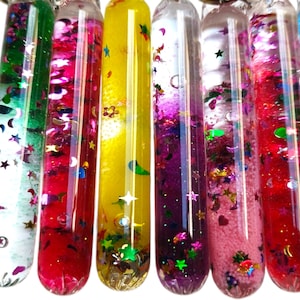 Könnte beinhalten: Acht durchsichtige Glasröhrchen gefüllt mit buntem Glitzer und Flüssigkeit. Die Röhrchen sind in einer Reihe angeordnet und alle gleich groß. Der Glitzer ist in verschiedenen Farben, darunter Rot, Grün, Blau, Lila und Pink. Die Flüssigkeit ist klar und die Röhrchen sind oben verschlossen.