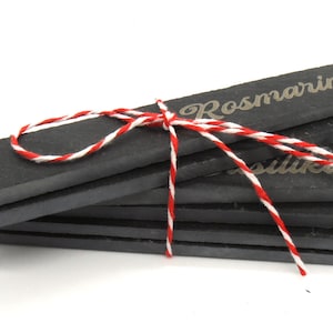 Op de afbeelding: Een set van zes zwarte leisteen plantenstickers gebonden met rood en wit touw. De stickers zijn gegraveerd met de woorden "Rosmarin" en "Salbei".