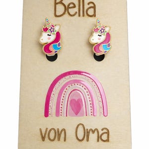 Könnte beinhalten: Holz-Display-Board mit zwei Einhorn-Ohrringen und einem rosa Regenbogen-Design. Das Board trägt den Namen "Bella" und den Schriftzug "von Oma" in braunen Buchstaben. Die Ohrringe haben goldfarbene Verzierungen.
