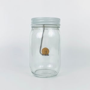 May include: A clear glass jar with a white lid and a metal hook inside. A small, round, brown cork is hanging from the hook.